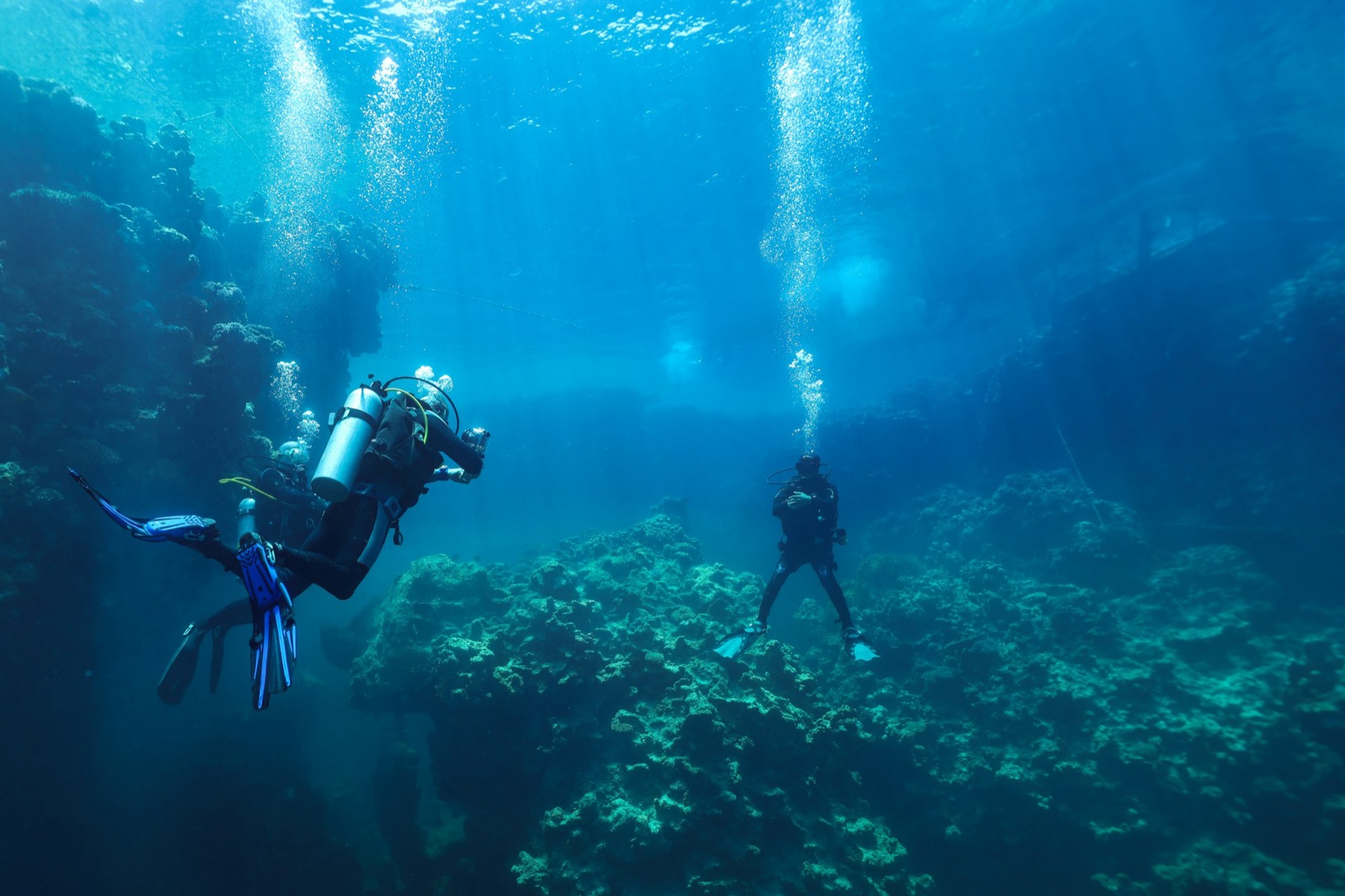 Scuba divers swim over a coral reef while bubbles rise through clear blue water