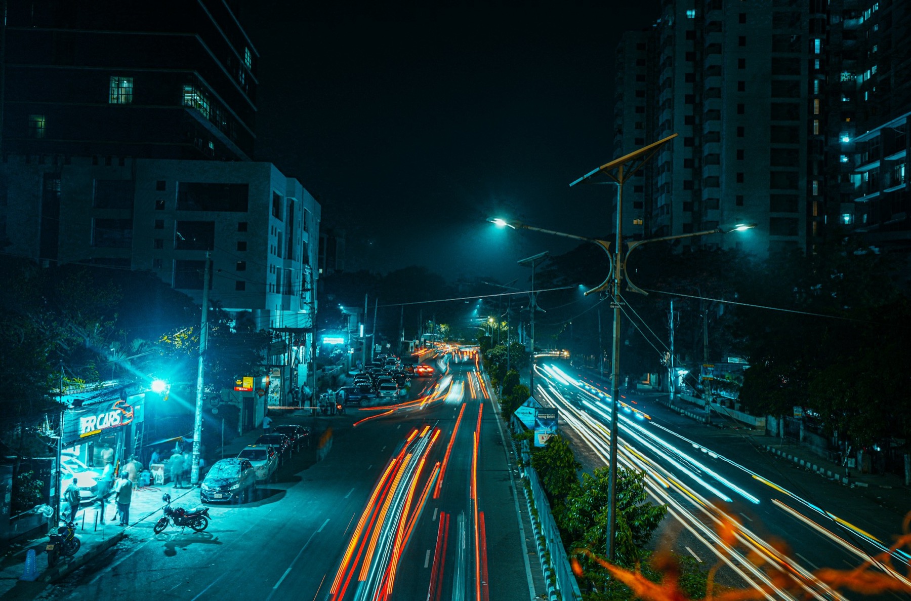A long-exposure night street in Dhaka with light trails from cars and teal-lit buildings