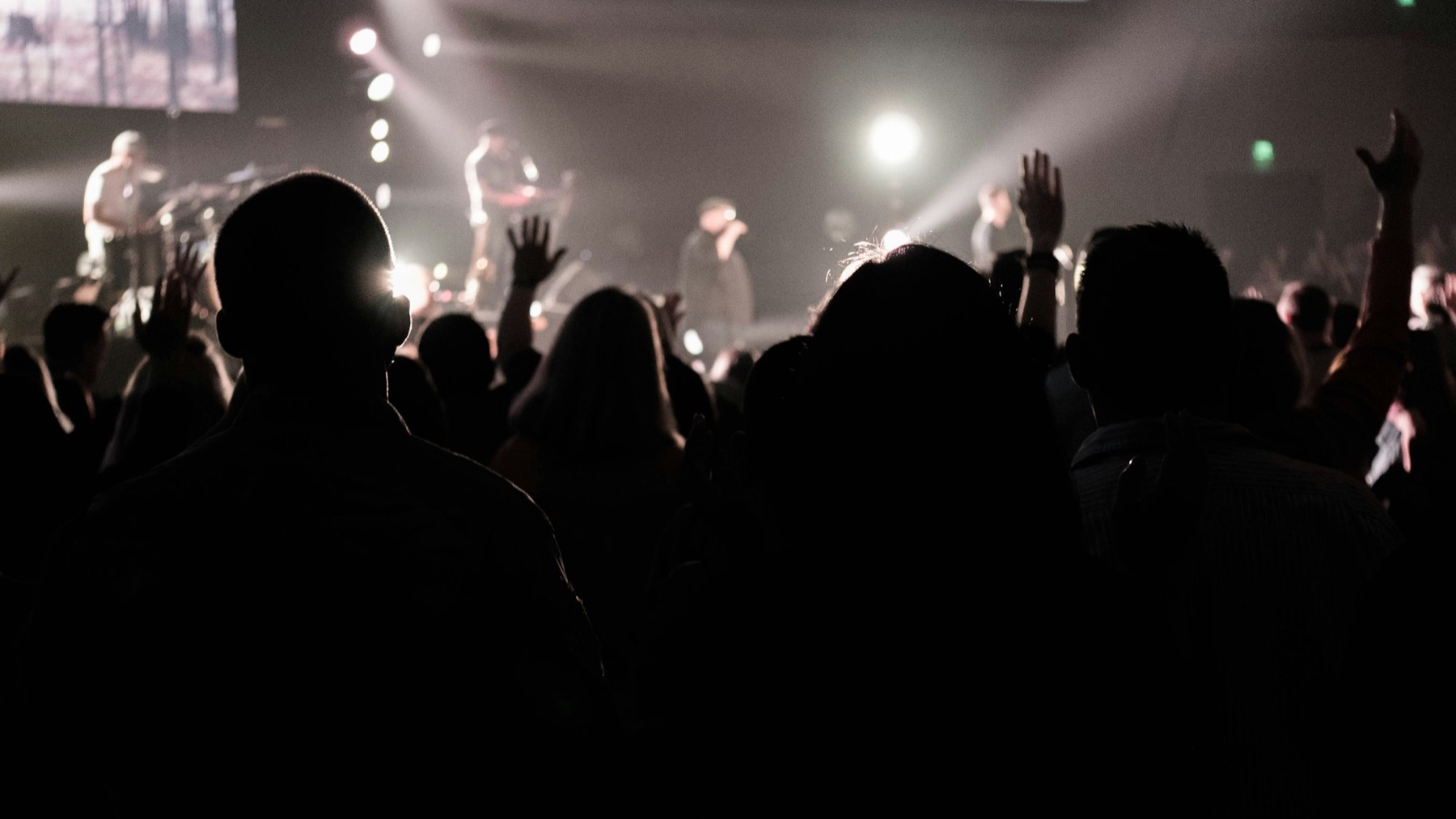 A crowd silhouetted against stage lights, hands raised, watching a live band perform
